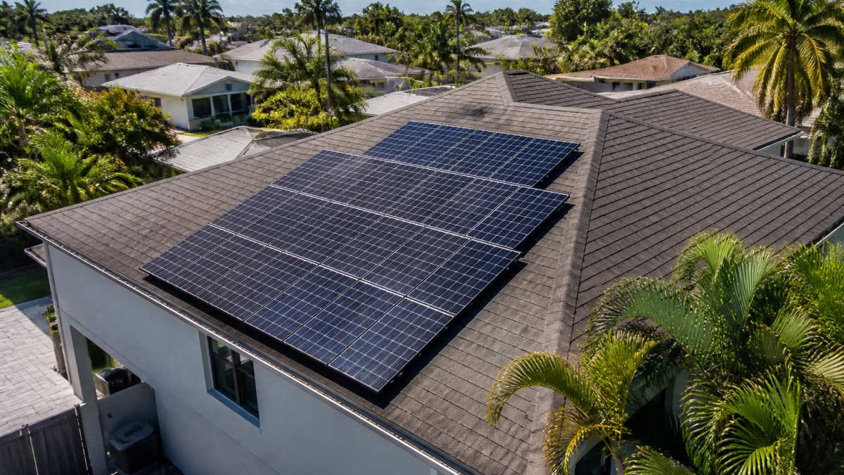 Aerial view of a suburban Florida home with rooftop solar panels gleaming in bright afternoon sunlight, palm trees visible in the background